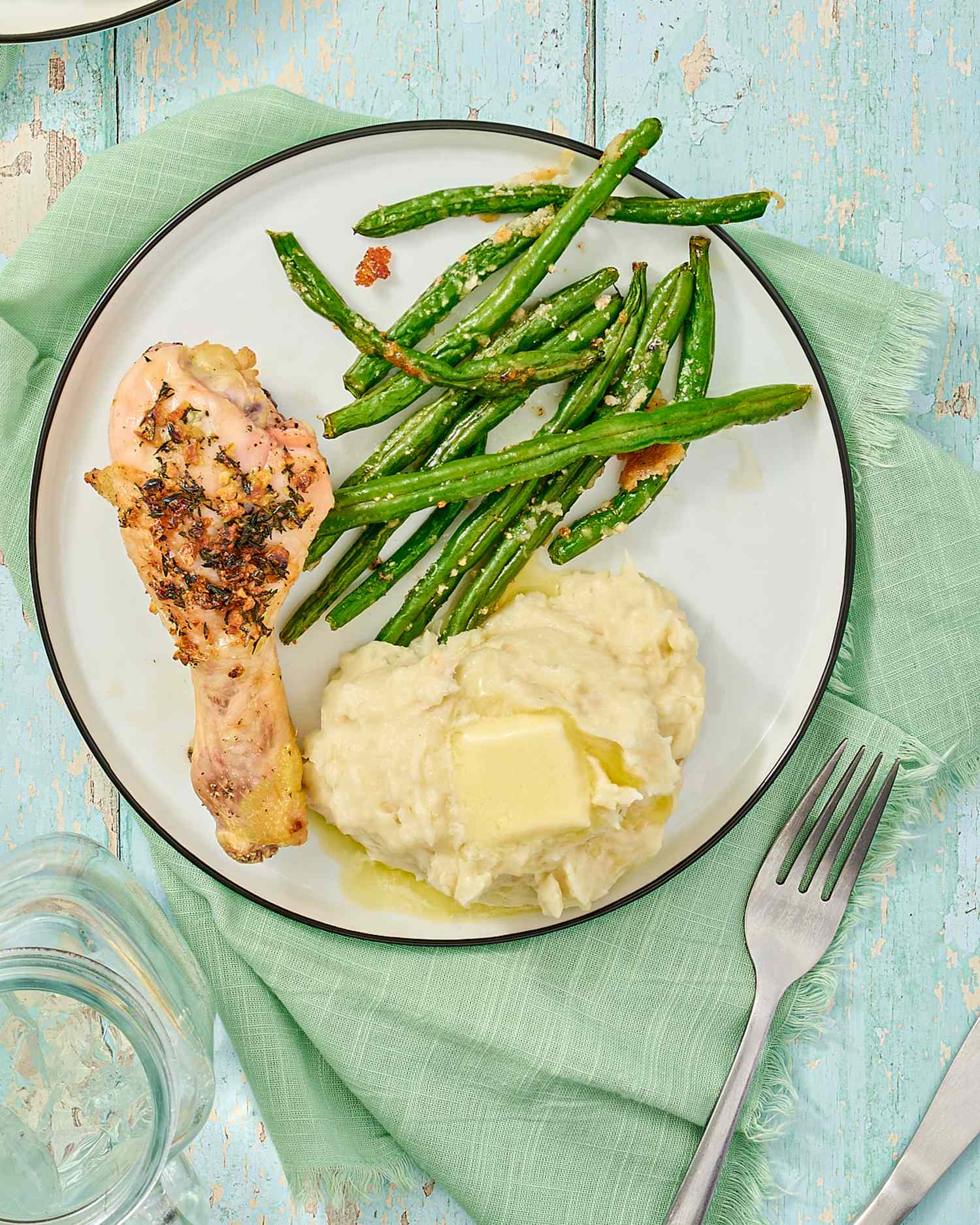 Plate with a chicken drumstick mashed potatoes with butter and green beans on a lightcolored table