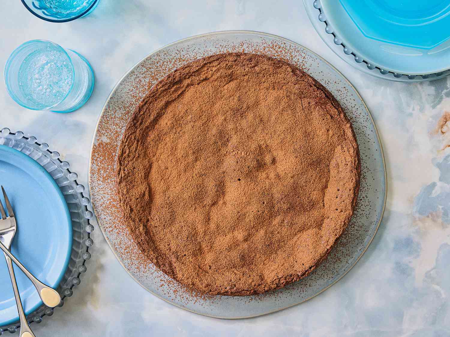 Flourless chocolate cake on light blue marbled surface, around the sides are various blue plates, blue cups and silver forks 