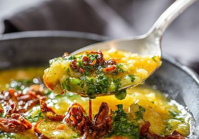 Closeup of a spoon lifting a portion of a dish with cooked rice garnished with herbs and fried shallots from a bowl