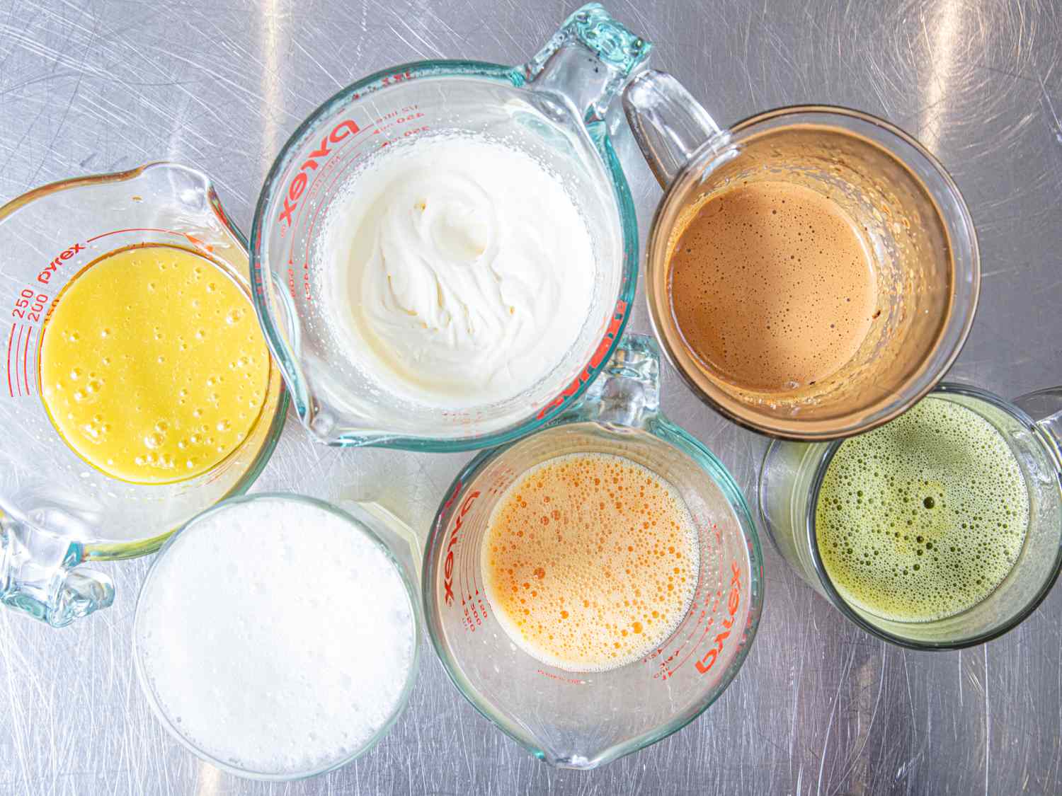 Six different beverages including milky and frothy textures displayed in measuring cups on a metal surface