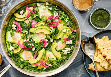 Pot of shakshuka on grey stone surface, with coffee cup, and side of pita. 