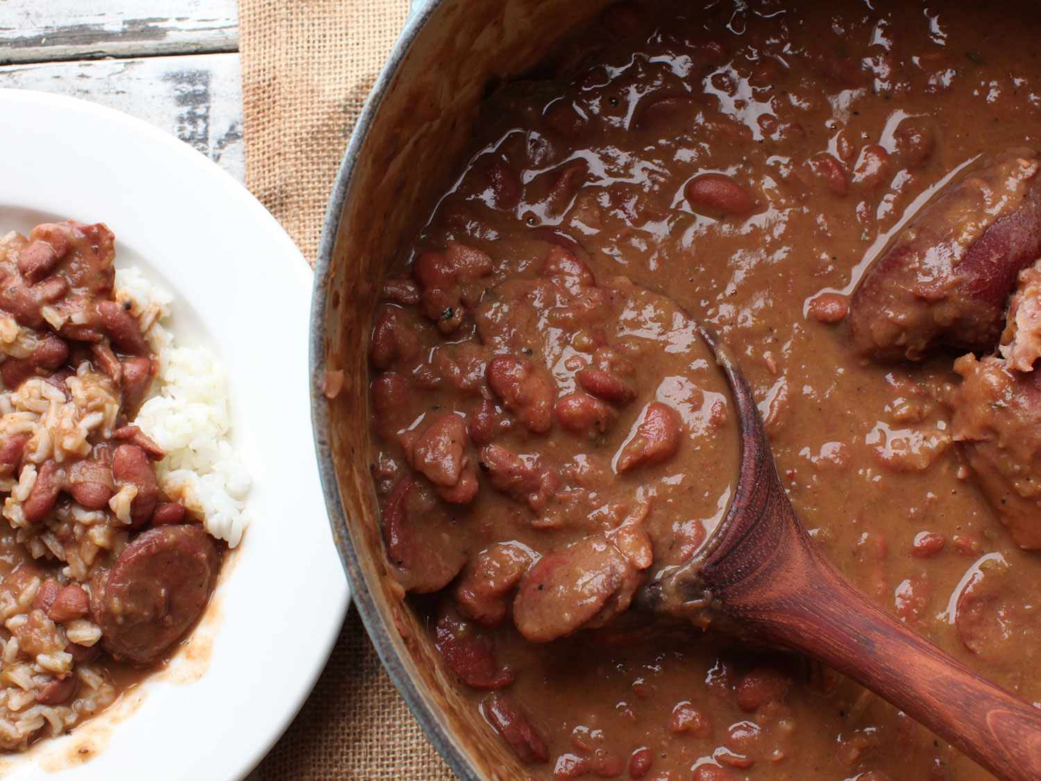 A Dutch oven with cooked New Orleans style red beans, and on the left is a shallow bowl of red beans and rice with slices of sausage. 