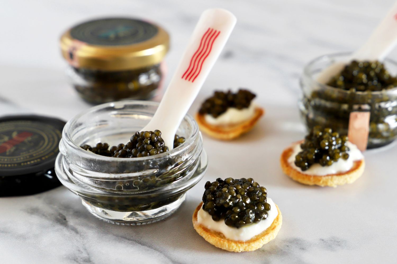 a jar of Caviar Co royal white sturgeon caviar next to a blini topped with crème fraîche and a scoop of caviar