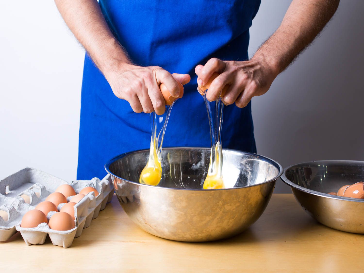 A person cracking open two eggs simultaneously into a metal bowl, with an egg in each hand.