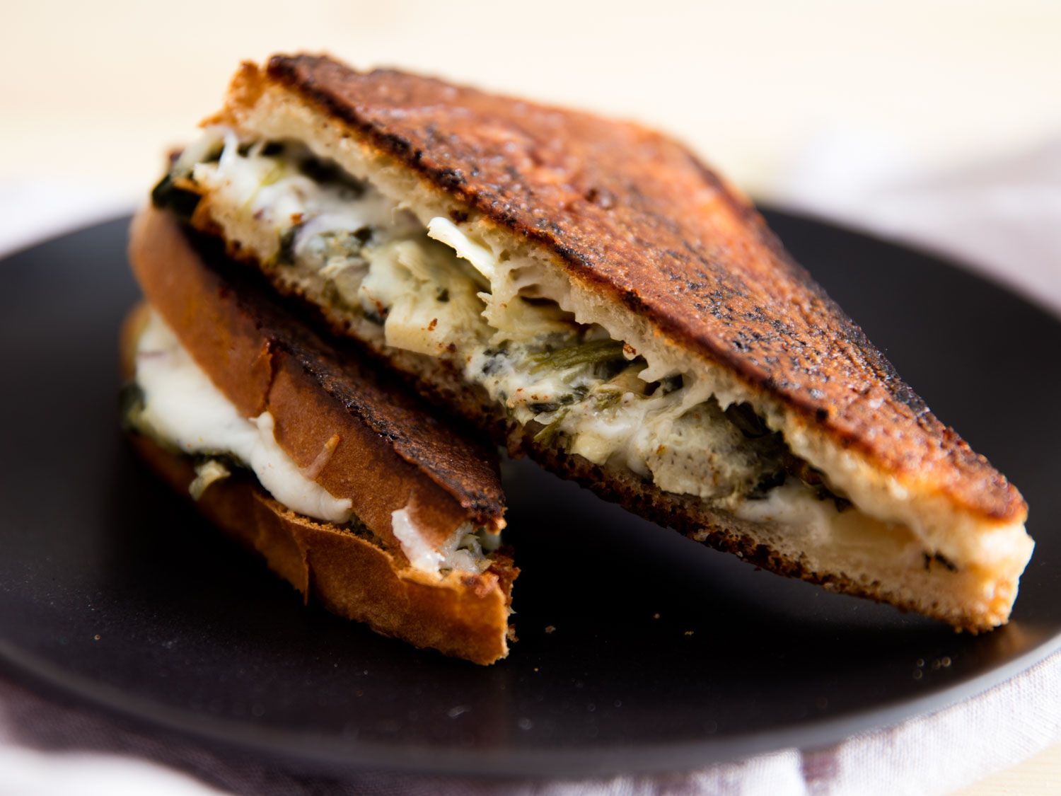 A cut in half grilled cheese sandwich, filled with artichokes and greens on a black plate.