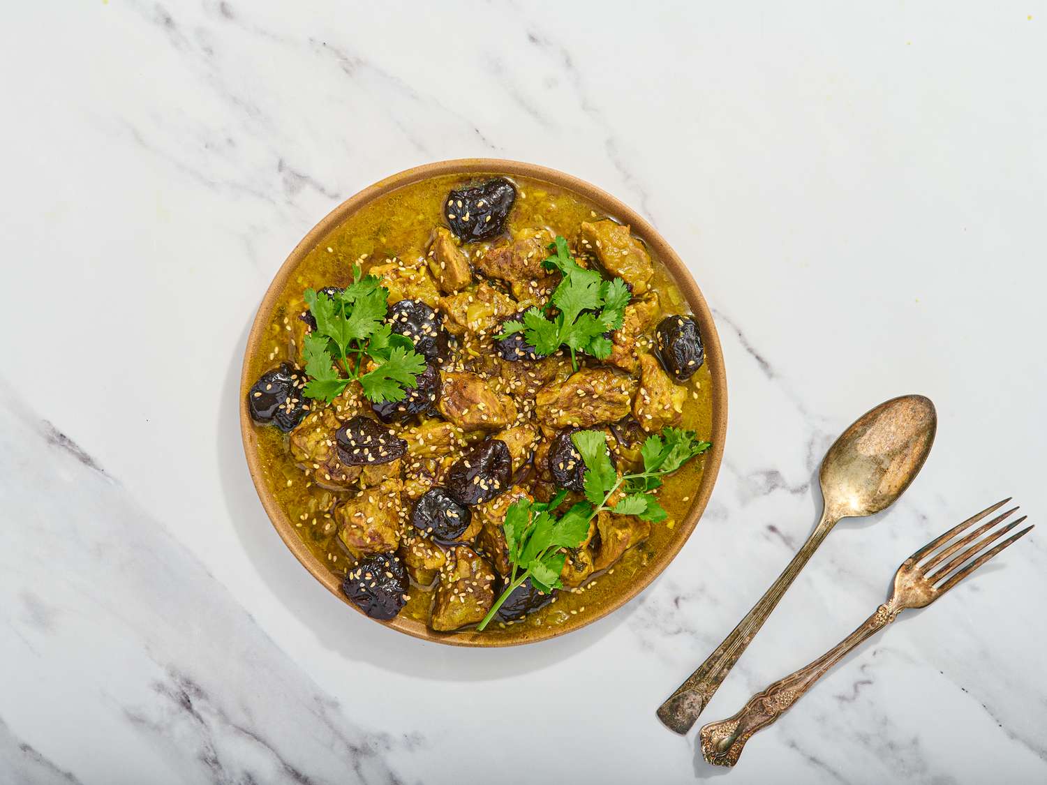 Tagine in a stone bowl garnished with prunes and cilantro, and rustic cutlery 