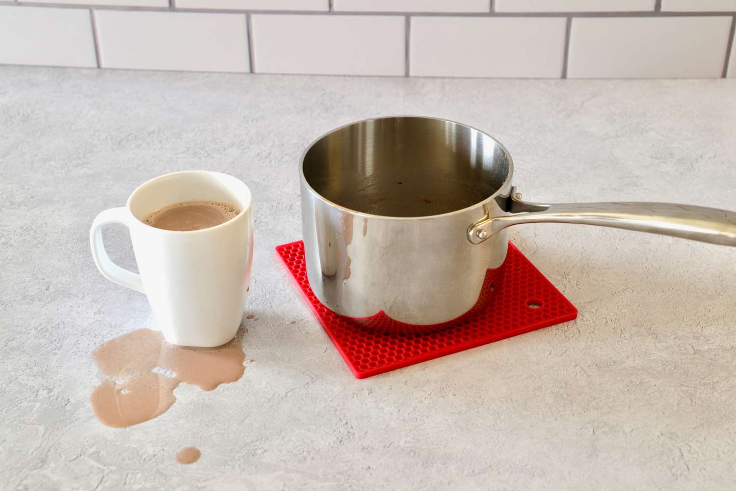 A small saucepan beside a mug of hot cocoa with some hot cocoa spilled on the counter