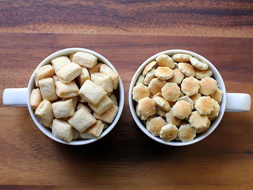 Overhead view of two mugs. One is piled high with homemade, square-shaped oyster crackers. The other contains hexagonal store-bought crackers.