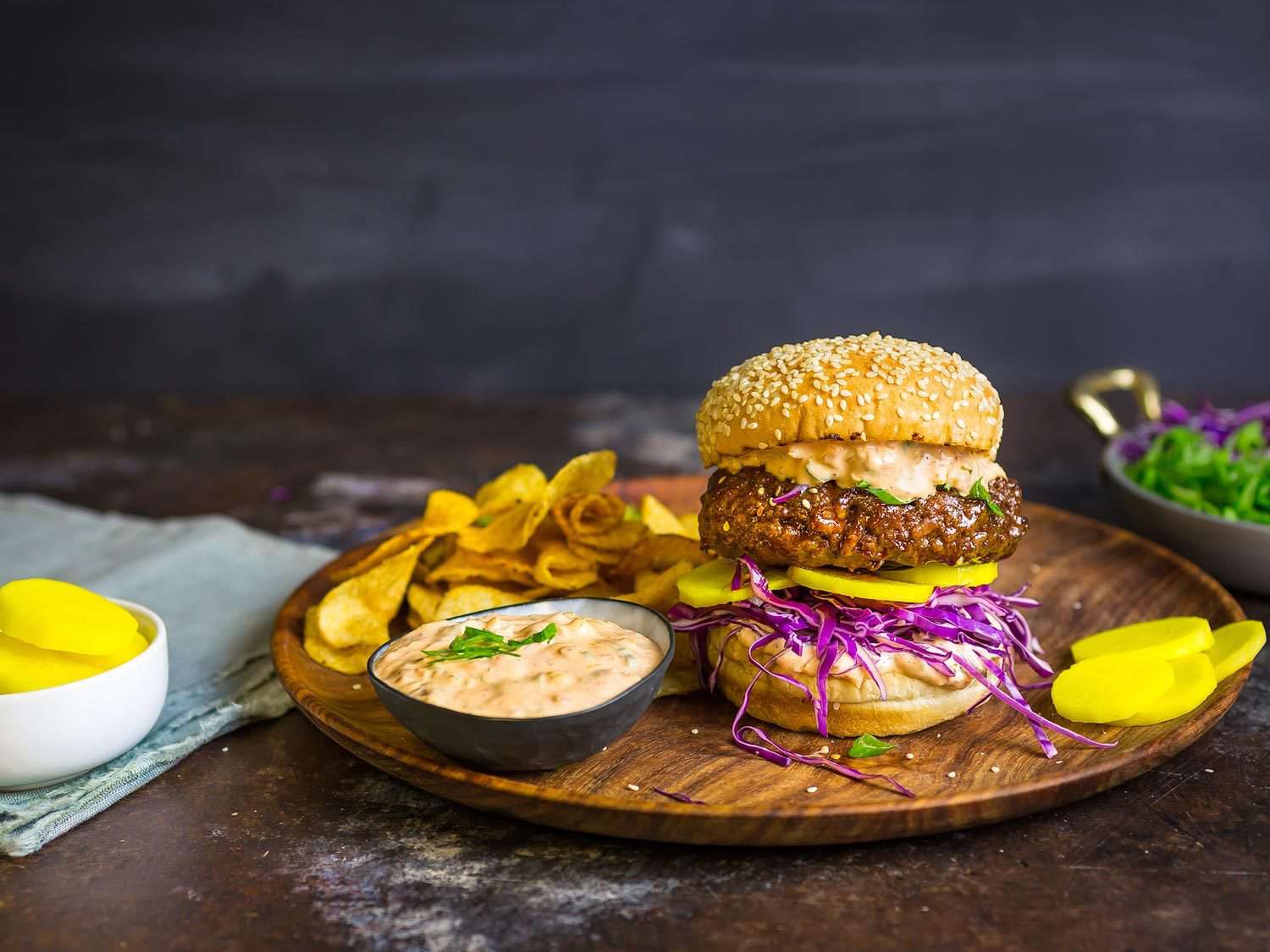 Burger on a wooden serving board with sauce and chips.