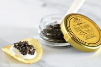 An open jar of beluga hybrid caviar next to a potato chip topped with crème fraîche and a scoop of caviar