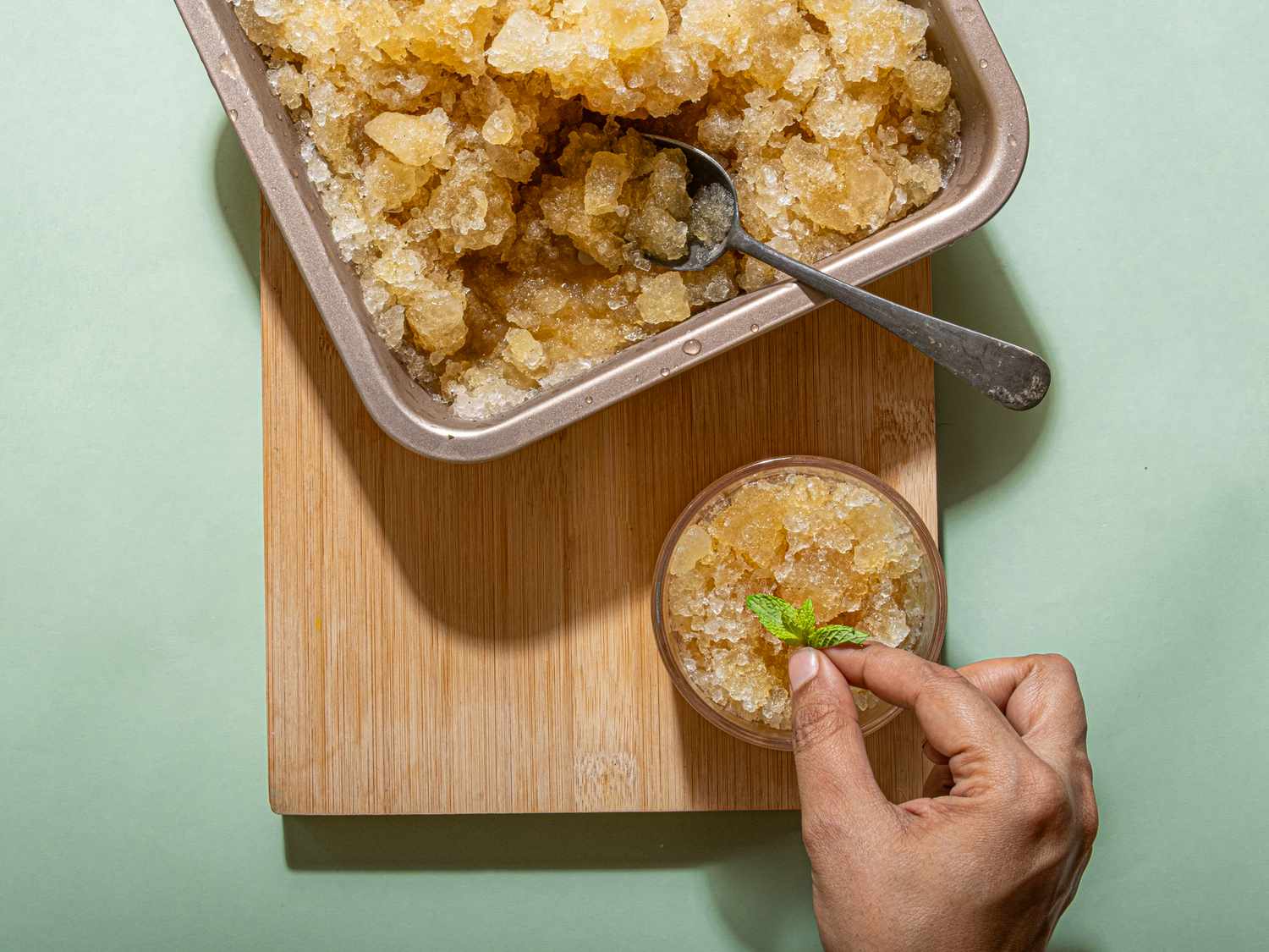 Granita in a tray and a glass being garnished with a mint leaf on a wooden block