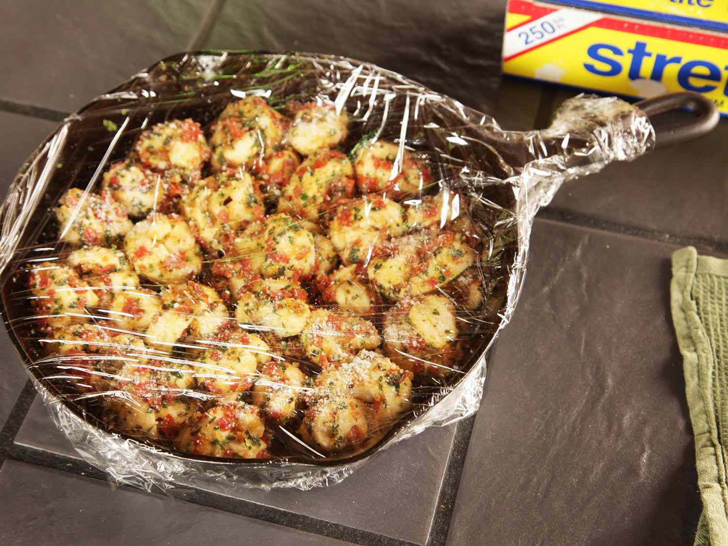 A plastic wrap-covered cast iron pan filled with proofing pepperoni garlic knots