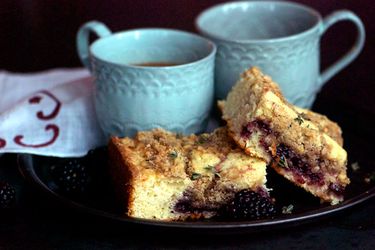 Two slices of blackberry thyme crumb cake, served on a black plate with two mugs of coffee.