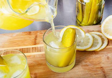 Glass of lemonade with a pickle spear lemon slices and a mason jar with pickles nearby on a wooden surface