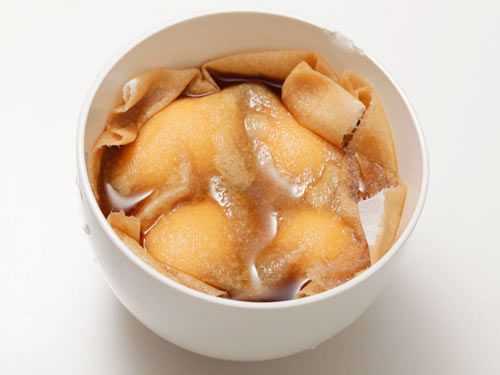 Four soft-boiled eggs in a soy sauce marinade in a white bowl, covered with a paper towel to force the marinade over the tops of the eggs.