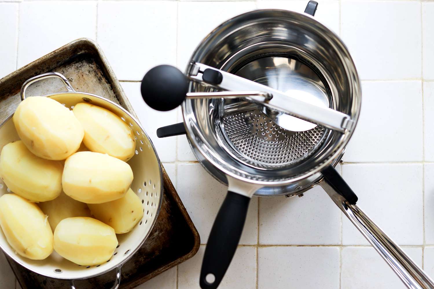 A food mill with boiled potatoes in a steamer basket on a white tile surface