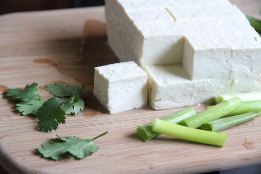 Cilantro leaves, cubes of tofu, and length of green onion on a wooden cutting board.