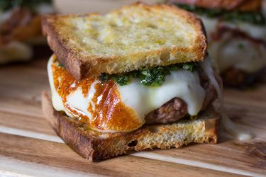 Three asado burgers on a wood cutting board, each made on sliced bread and topped with melted cheese and chimichurri. 