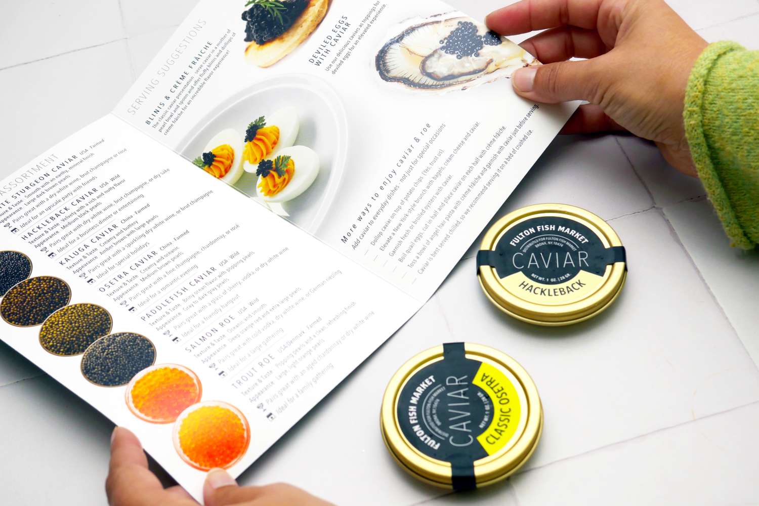 Hands holding a menu with caviar dishes pictured next to two tins of caviar on a table