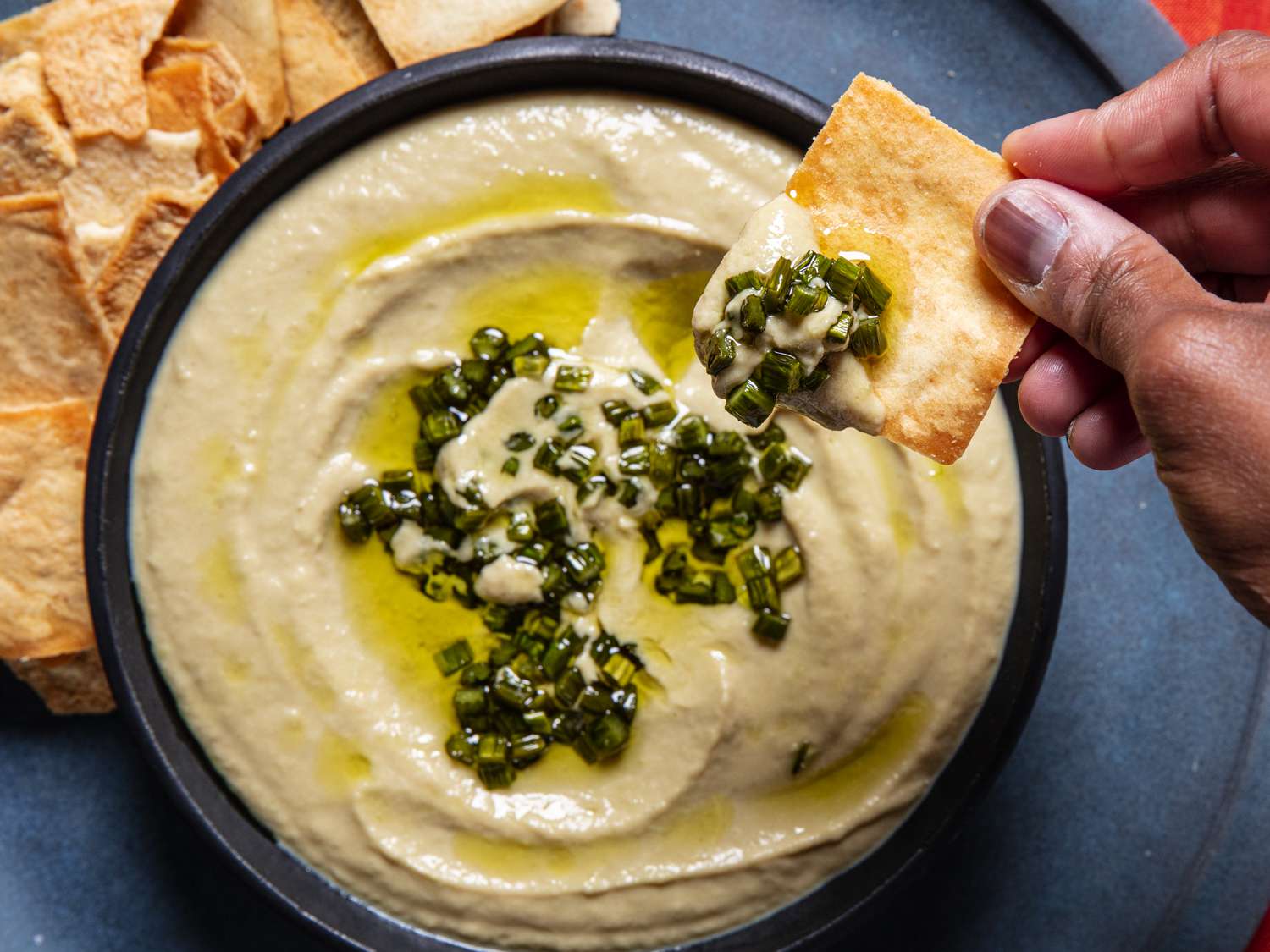 A hand lifting a pita chip with garlic scape hummus on it over a bowl of hummus