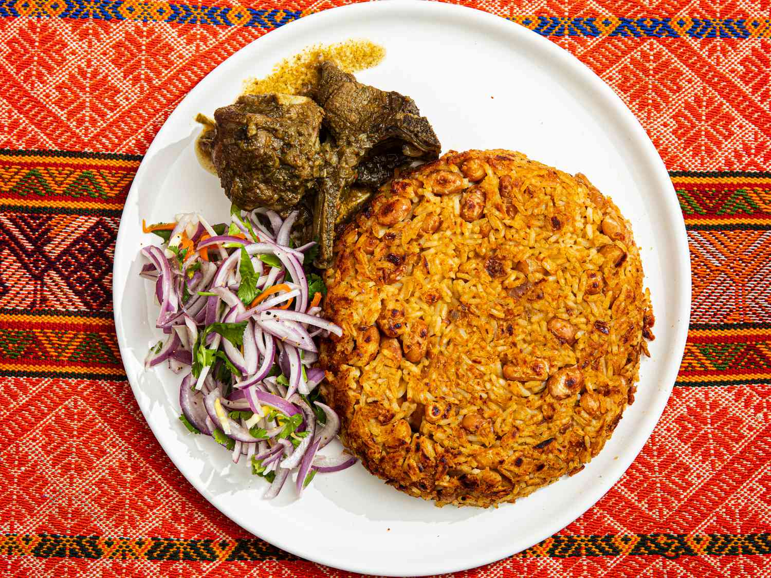 Overhead view of finished tacu tacu on a white plate with salad and leftover stew on a traditional Peruvian fabric 