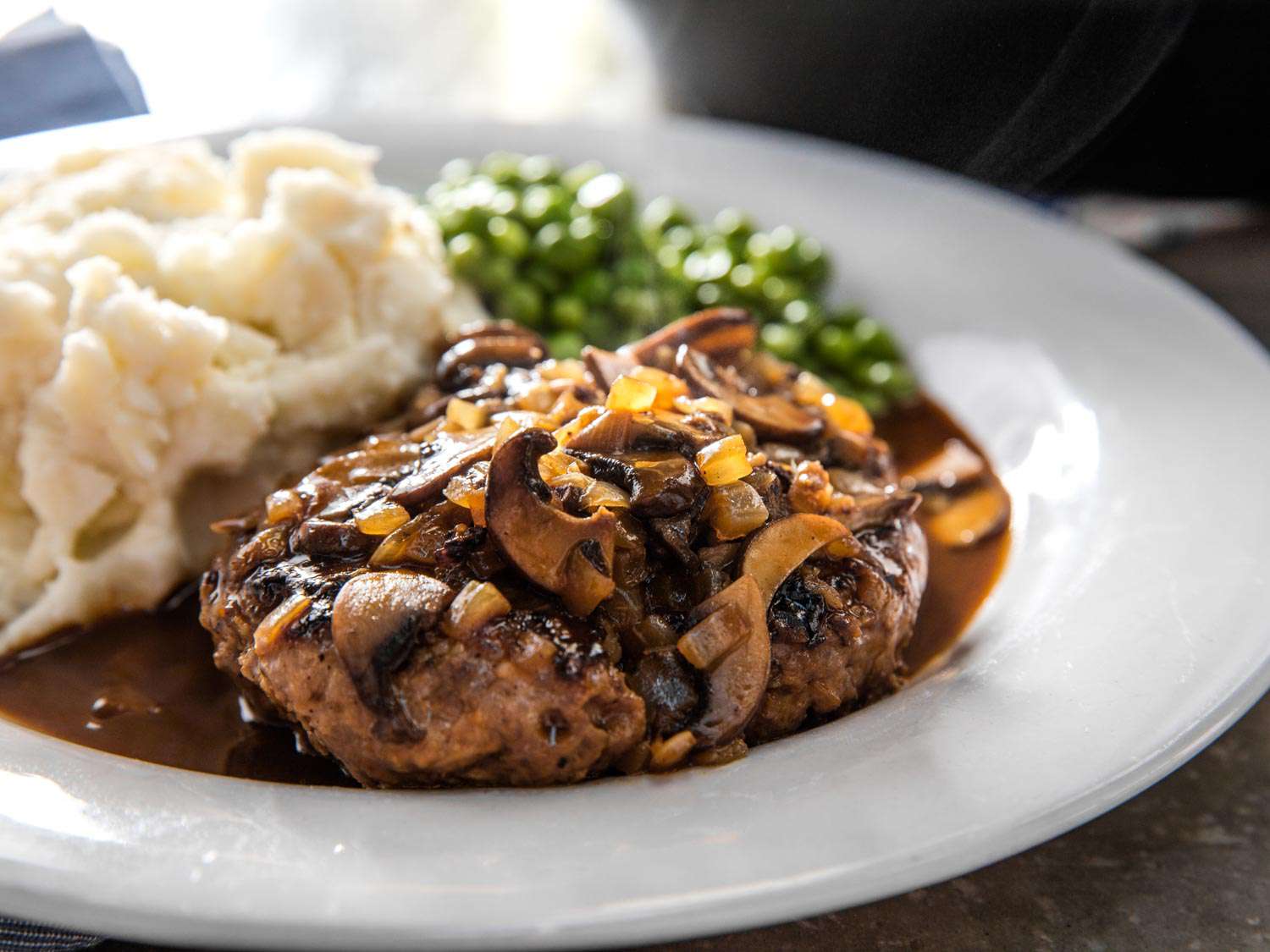 Salisbury steak smothered in mushroom onion gravy, plated with mashed potatoes and peas