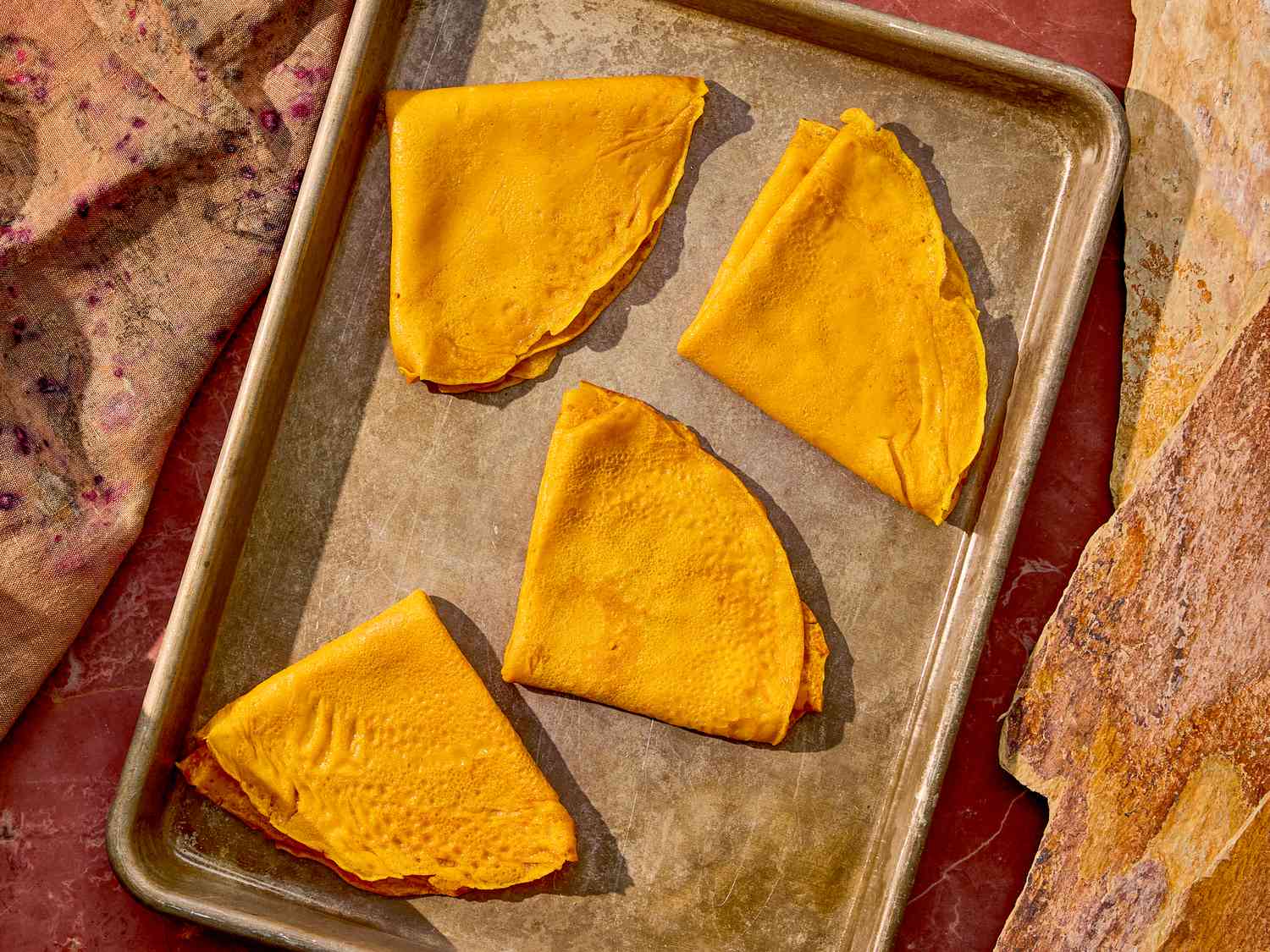 Four folded crepes on a baking tray, surrounded by fabric and bread on a surface