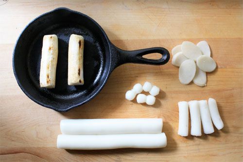 Korean rice cakes in different shapes: two cylindrical blocks browned in a cast iron skillet, while other shapes are uncooked, including rice cakes cut into thin slices and formed into balls. 