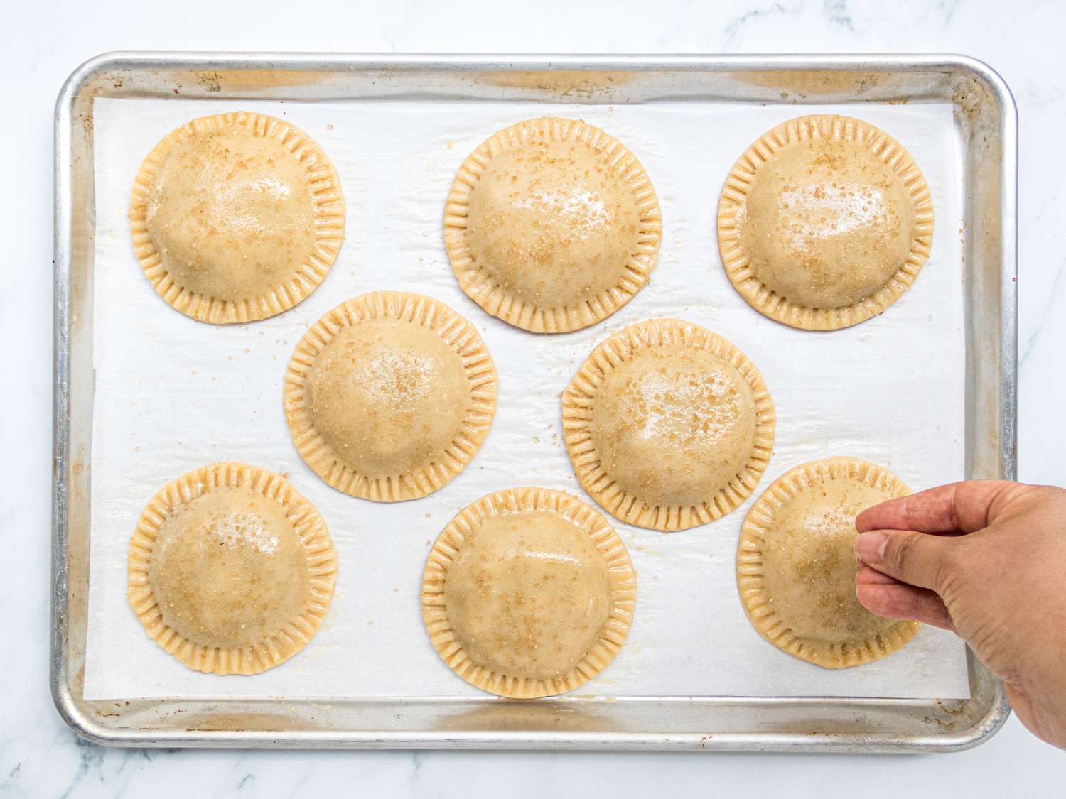 Handpies assembled on a baking sheet, with turbinado sugar being sprinkled on 