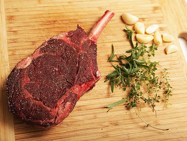 Seasoned raw bison ribeye steak, fresh rosemary, thyme, and whole garlic cloves on wooden surface.
