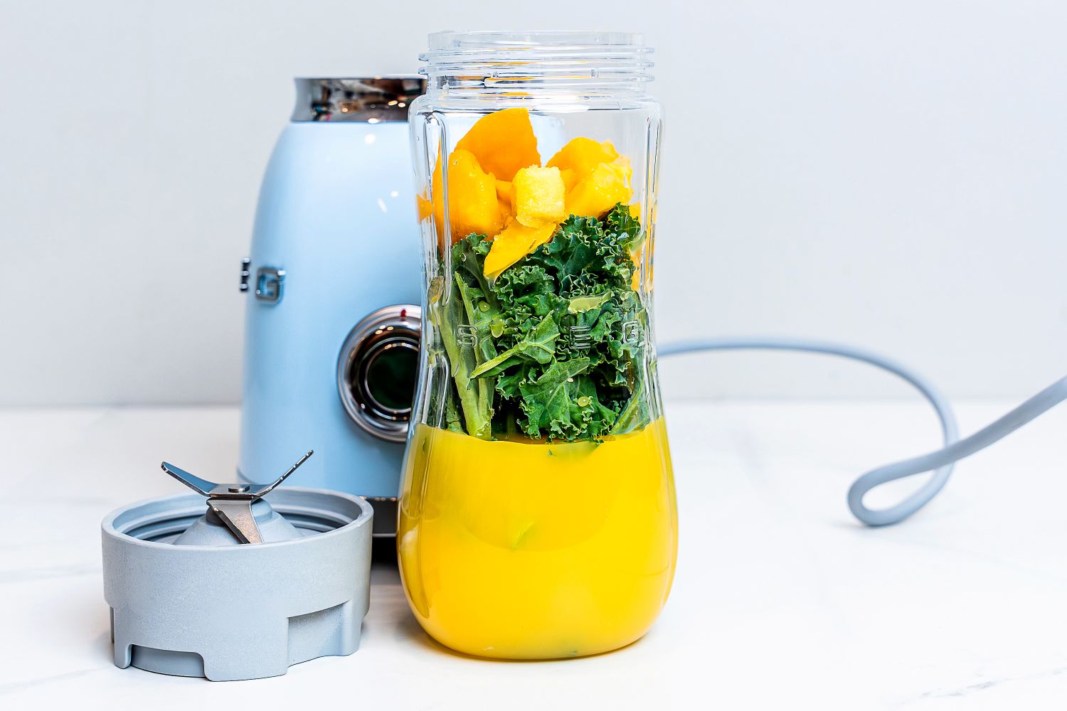 Blender jar filled with orange liquid, kale, and fruit pieces next to a blender machine
