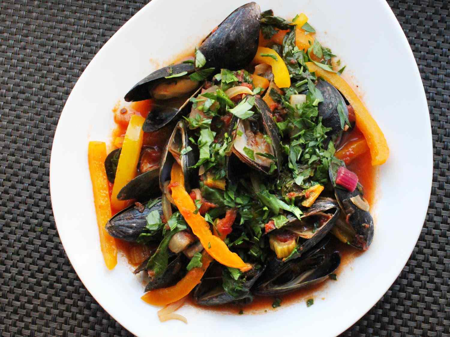Overhead of a white bowl filled with steamed mussels with peppers, chard, and tomatoes.