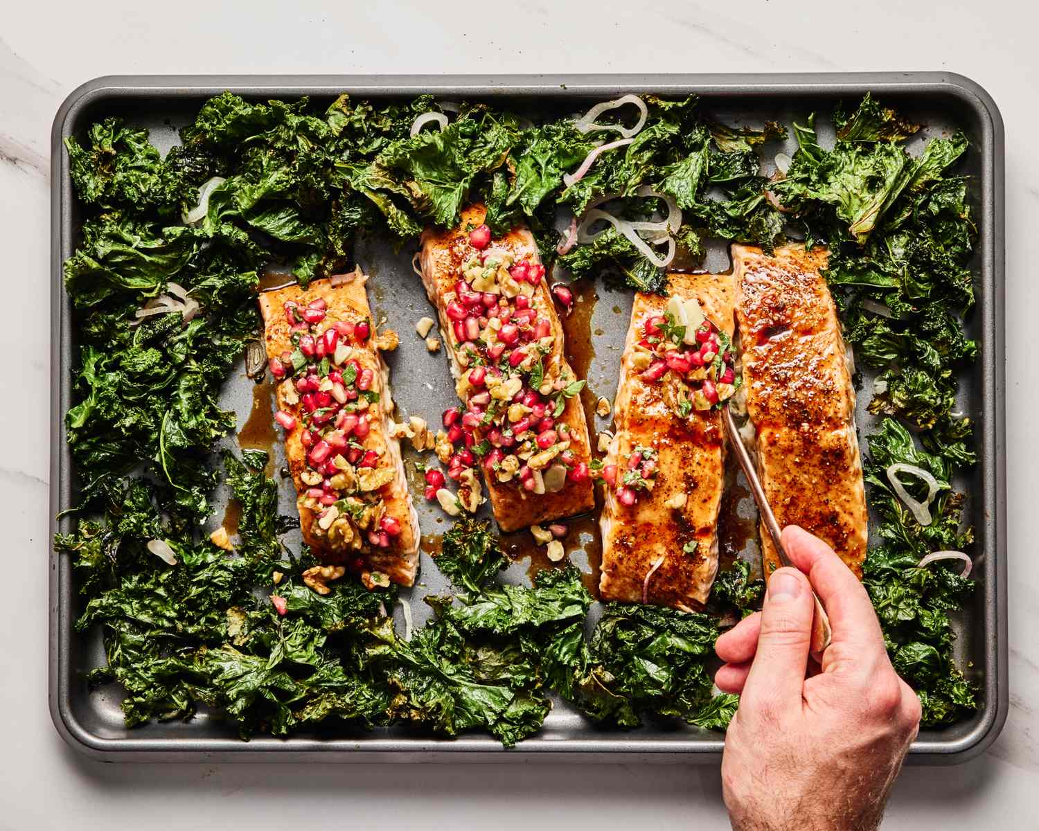 A tray of pomegranateglazed salmon with kale a hand places a utensil near one of the salmon pieces