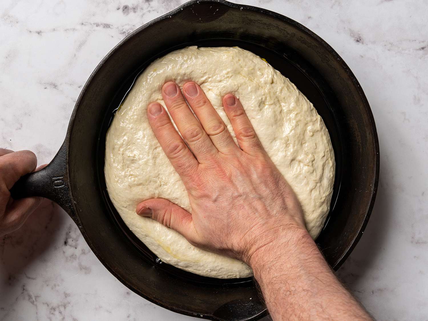 Hands pressing focaccia dough into an oiled cast iron skillet