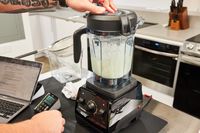 Hand resting on top of a Vitamix 7500 Blender blending ice and juice