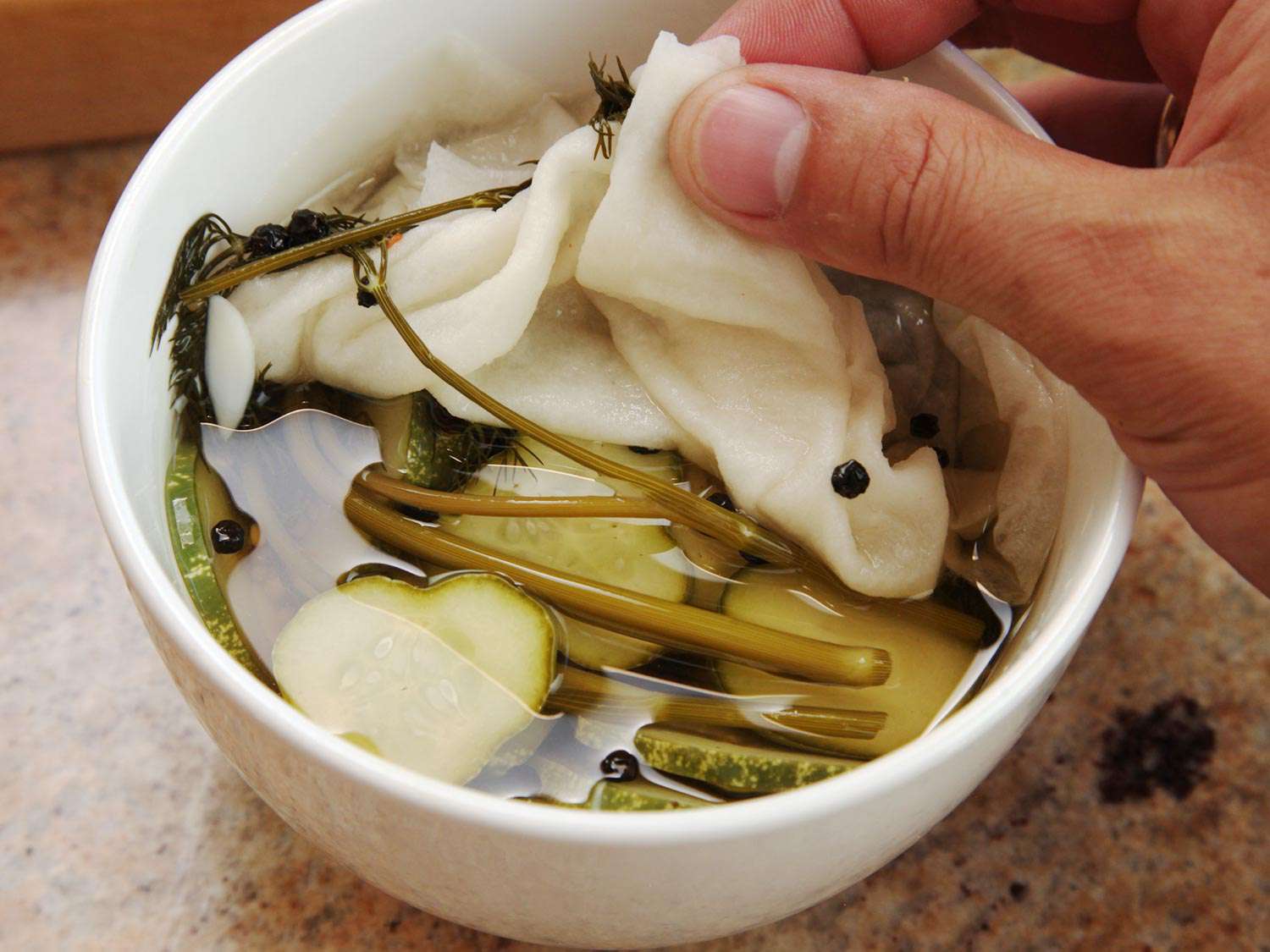 The ingredients for a quick pickle in a white bowl: sliced cucumbers, peppercorns, and herbs.
