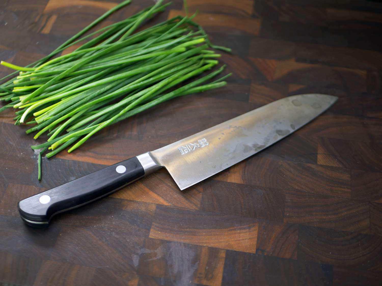 a carbon steel santoku on a wooden cutting board with chives