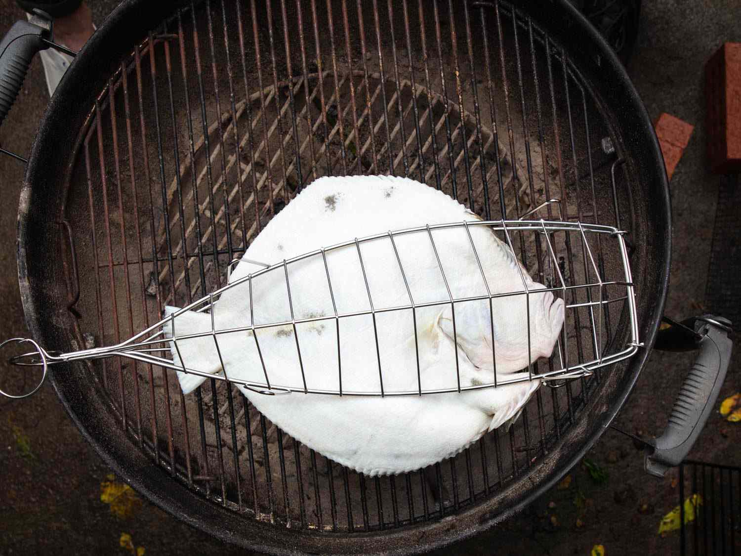 Overhead of turbot cooking in a fish-grilling cage on a charcoal grill, with white skin side facing up.