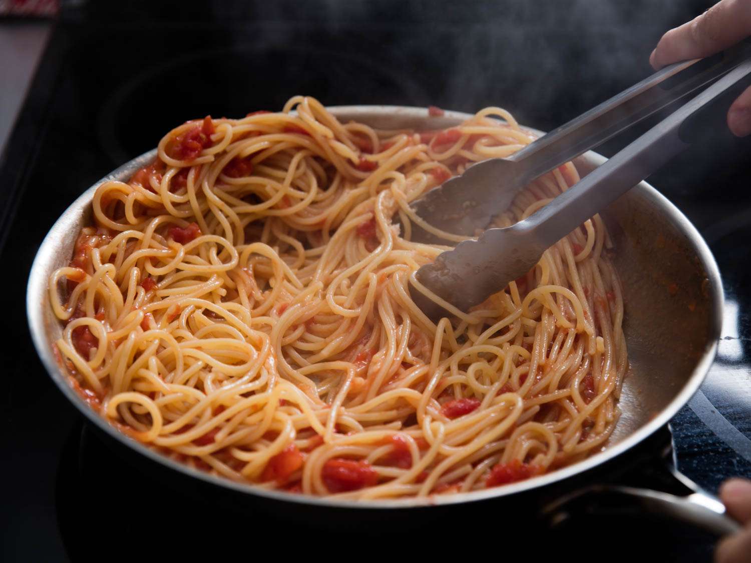 Pasta and sauce ingredients being stirred with tongs cooking in a skillet.
