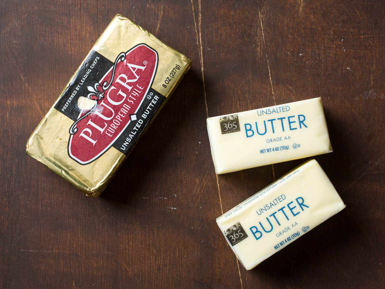 Overhead shot of three packages of butter (Plugra European-style butter and two packages of American-style unsalted butter) on a wooden background.