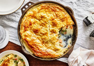 Baked Pot pie with one serving taken out of skillet and on a plate. Skillet is on a white tablecloth and has serving plates around it. 
