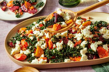 A platter containing kale salad with roasted root vegetables and crumbled cheese served with a wooden spoon