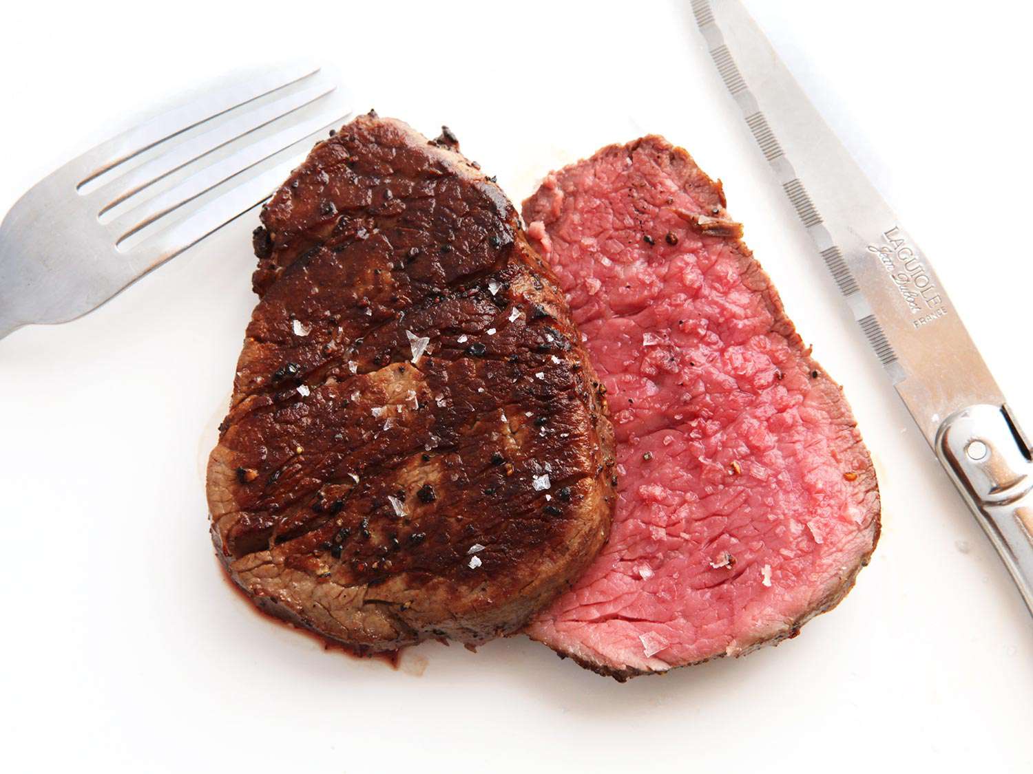 A steak cut in half resting on a white surface with a knife and fork off to the side.