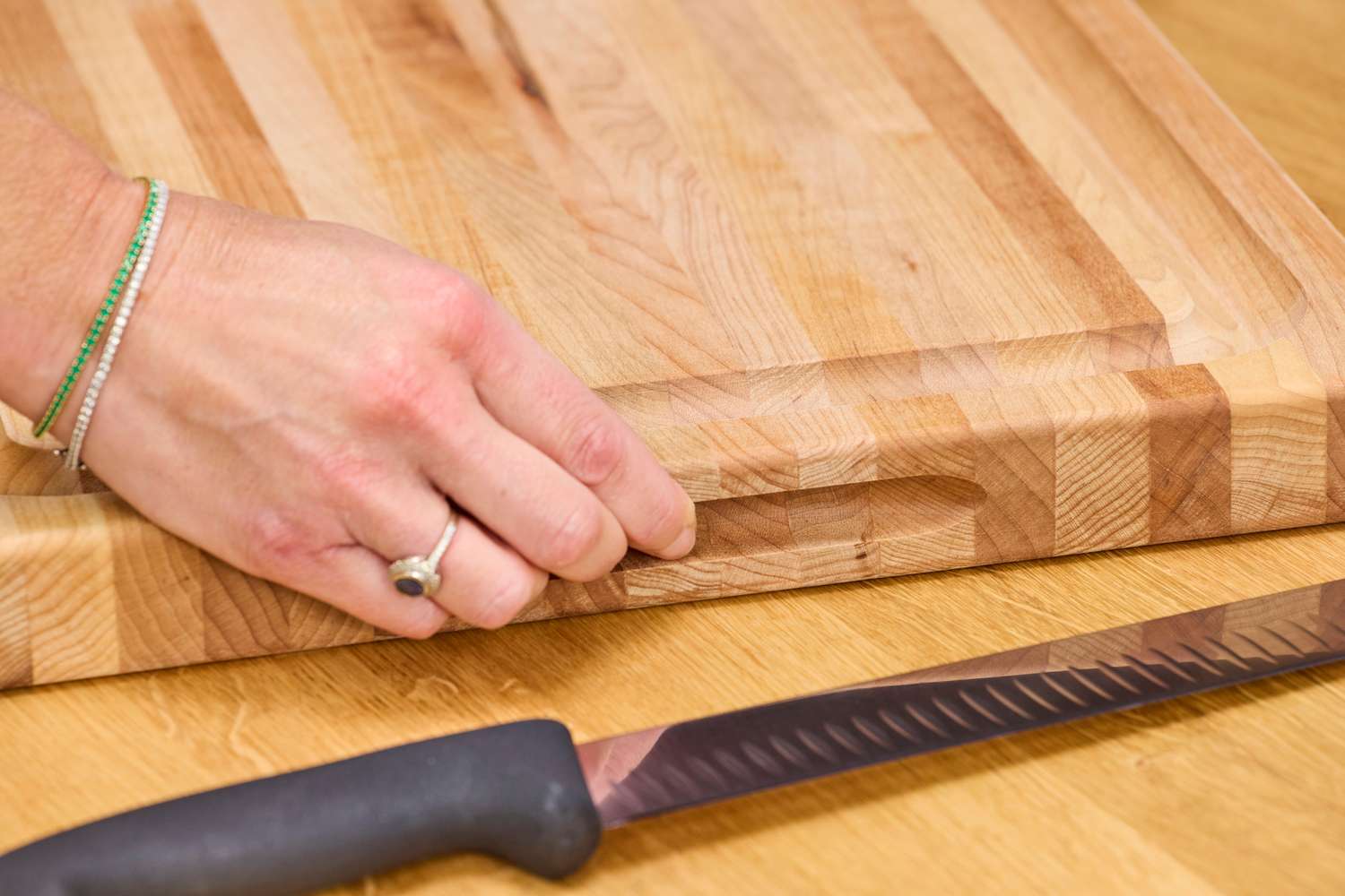 A hand adjusting a John Boos Reversible Maple Wood Cutting Board with Juice Groove on a table near a serrated kitchen knife
