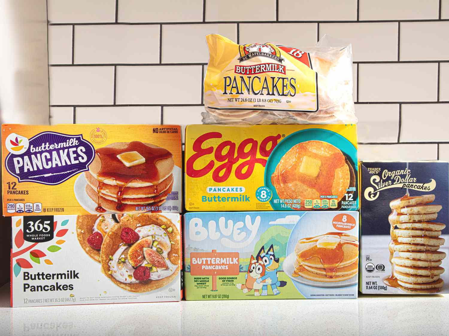 Five different brands of frozen pancakes displayed against a tile background including 365 Eggo and Buttermilk Pancakes packaging