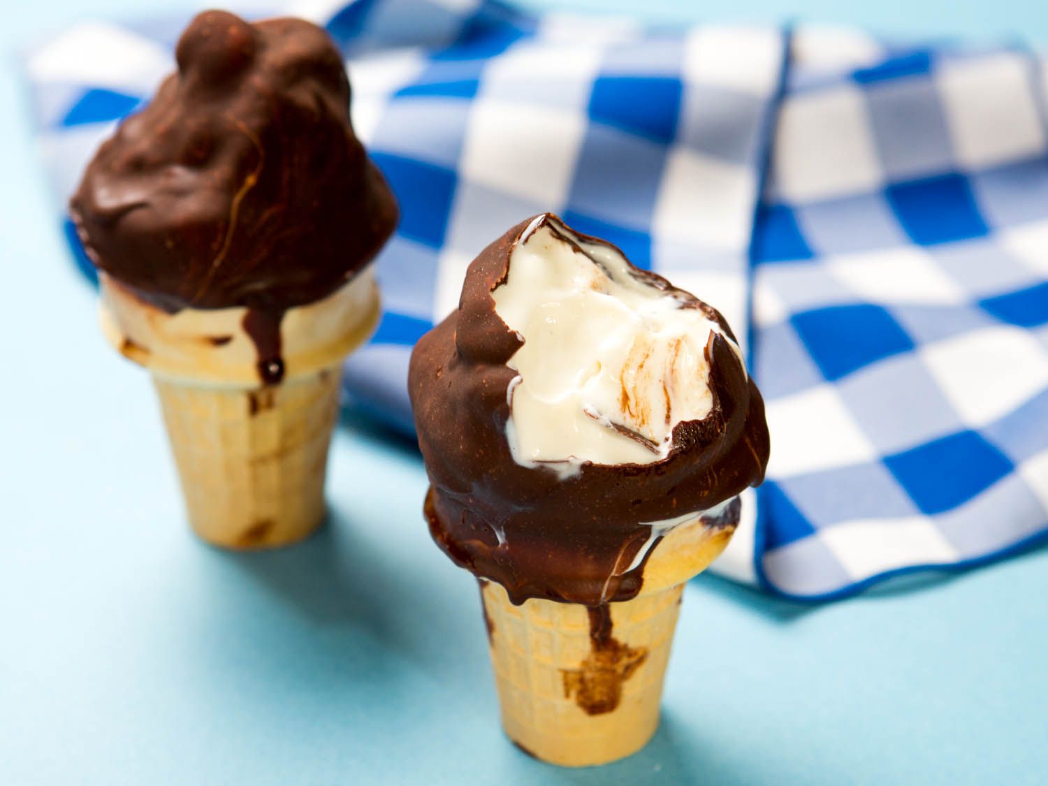 Two homemade chocolate-dipped soft-serve vanilla bean ice cream cones against a blue gingham napkin, a bite taken out of one.