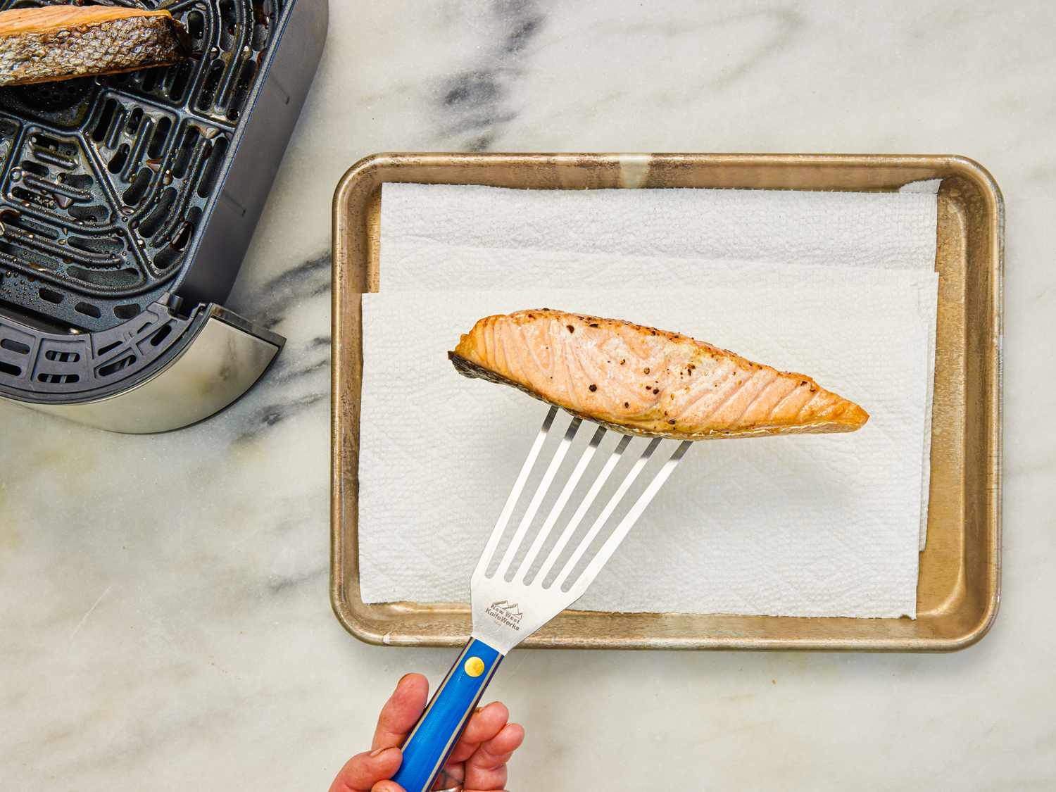 A finished piece of air fryer salmon is transferred to a paper-towel lined baking sheet.