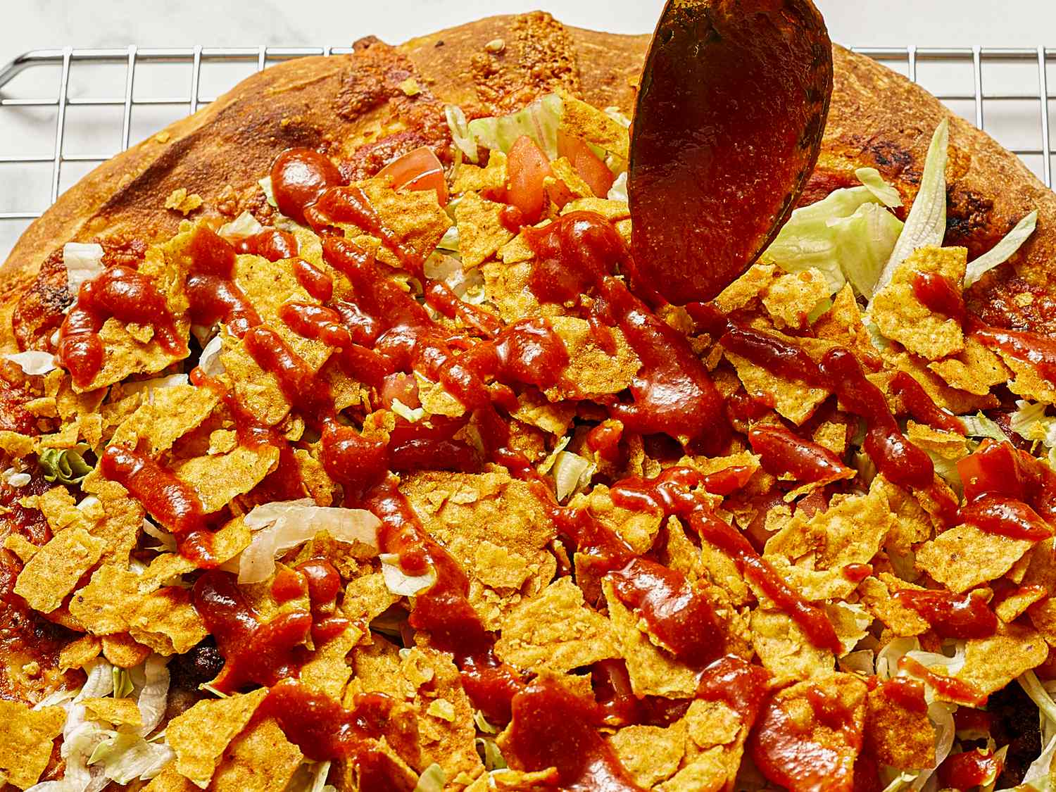 A pizza topped with lettuce crushed tortilla chips and a red sauce being applied by a spoon