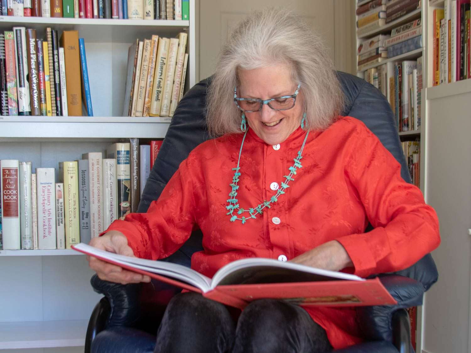 Cookbook collector Sue Jimenez, seated, reading from an open cookbook on her lap, with books on shelves behind her.