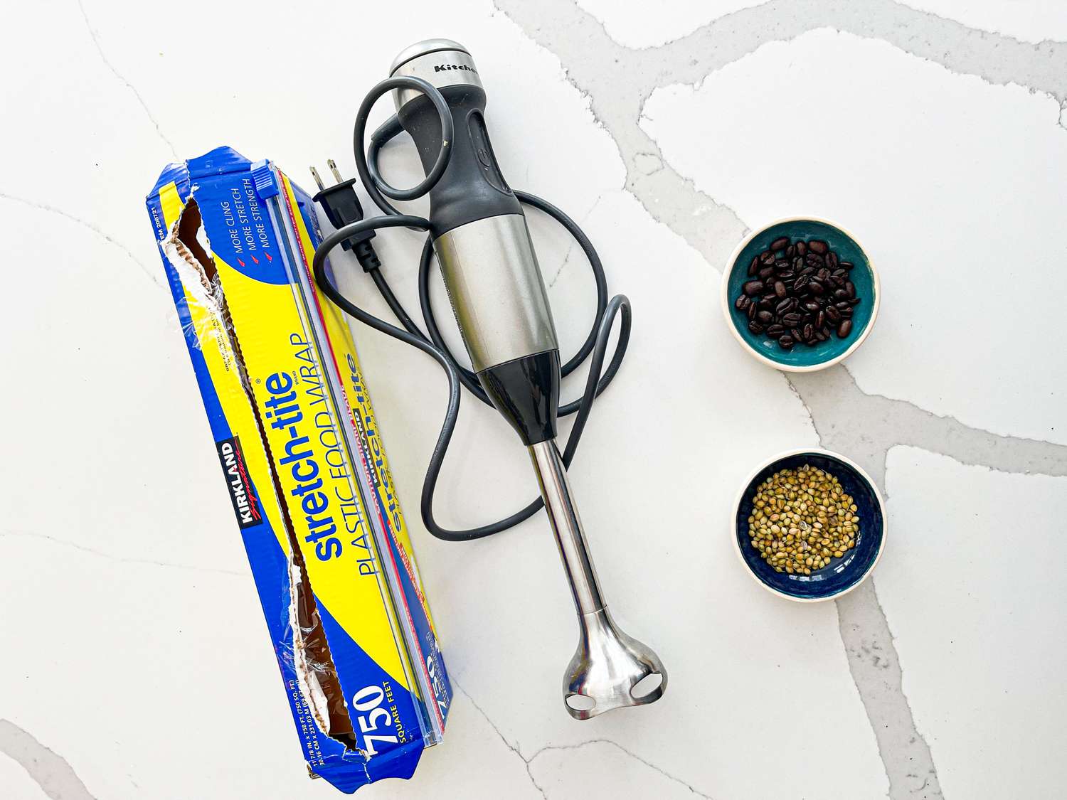A box of plastic wrap, an immersion blender, a small bowl of whole coffee beans, and a small bowl of whole coriander seeds.